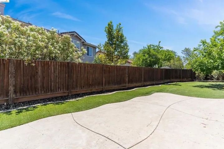 Expansive outdoor space featuring a patterned concrete patio, artificial turf, a wood privacy fence, and mature perimeter trees