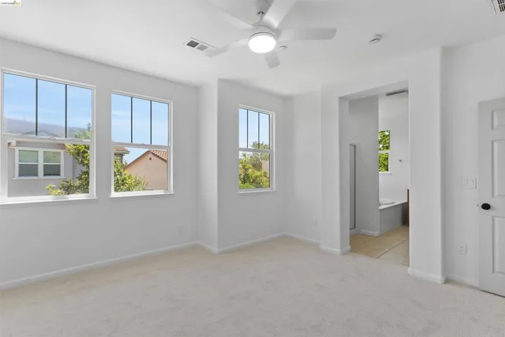 Bright room featuring three large windows, a ceiling fan with integrated lighting, and light-toned carpet flooring