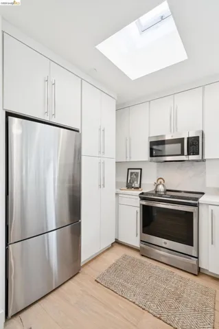 Kitchen featuring a skylight, white cabinetry, stainless steel appliances, light wood-finish flooring, and white countertops with a tiled backsplash
