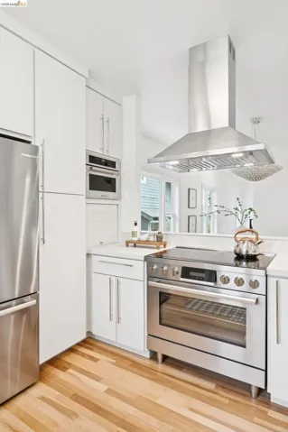 Modern kitchen featuring stainless steel appliances, white cabinetry, light countertops, a stainless steel range hood, and wood-finish flooring