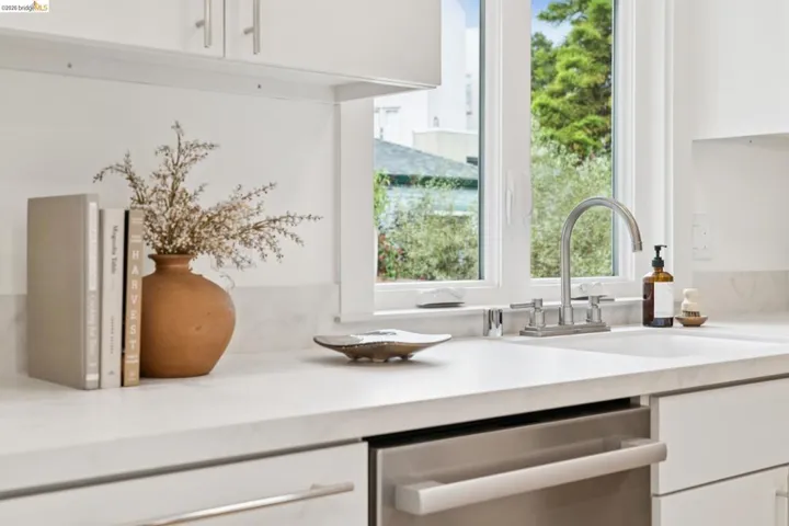 Kitchen featuring a white undermount sink, gooseneck faucet with separate hot and cold handles, stainless steel dishwasher, white cabinetry with brushed nickel pulls, and light-toned countertops