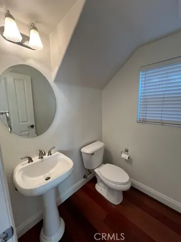 This well-appointed powder room features a pedestal sink, a framed round mirror, and a large window that fills the space with abundant natural light. Neutral wall tones and rich hardwood flooring create a warm, inviting atmosphere, while the clean white fixtures provide a timeless appeal. Thoughtfully designed and well maintained, this convenient main-level bathroom offers both functionality and style for guests and everyday living.
