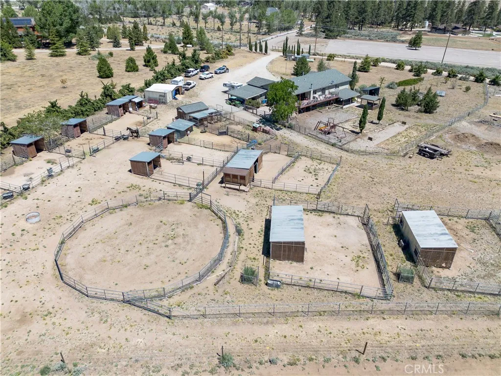 An elevated aerial perspective showcasing the equestrian facilities, highlighted by a large fenced round pen, multiple covered horse stalls, open turnouts, and a clear view of the surrounding property landscape.