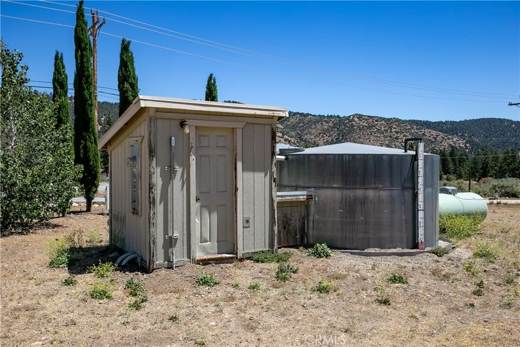 This view showcases a secure wooden pump house structure, a massive corrugated metal water storage tank, and an adjacent propane tank, all situated on the open, natural terrain with mountains in the distance.