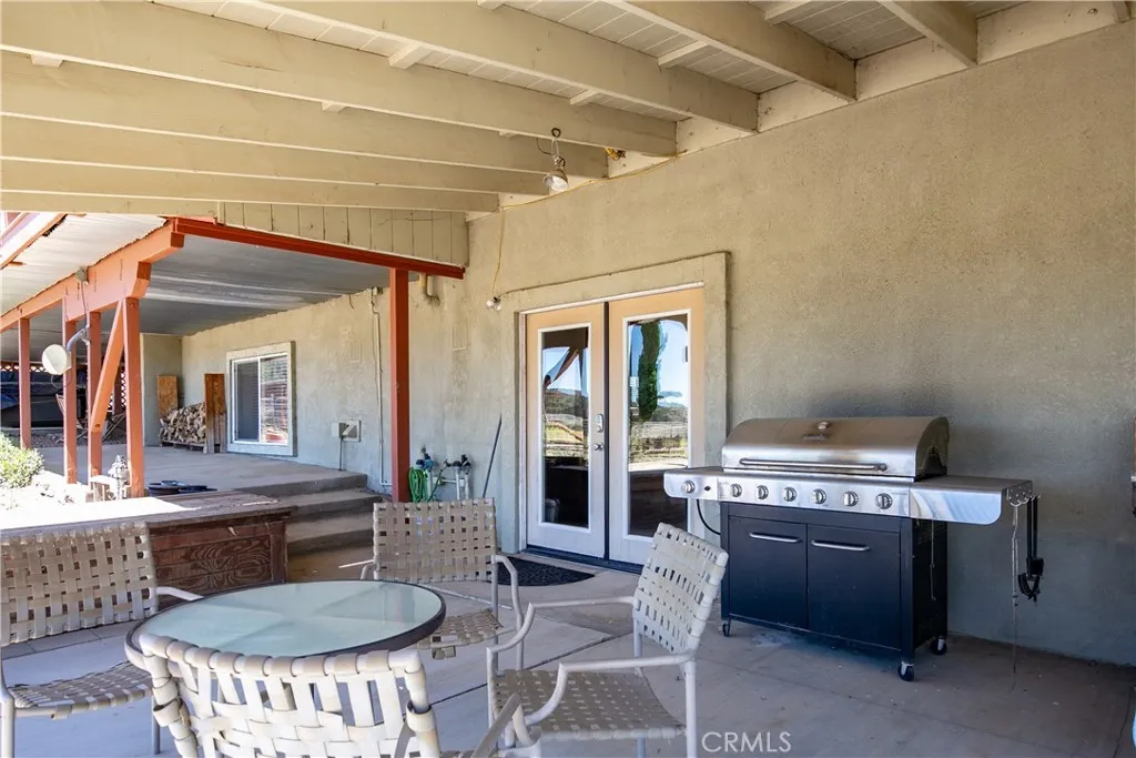 A welcoming ground-level perspective of the home's shaded outdoor living space. This covered concrete patio includes a cozy seating and dining layout, a premium stainless steel barbecue grill, and elegant French doors that seamlessly connect the indoor and outdoor entertaining areas.