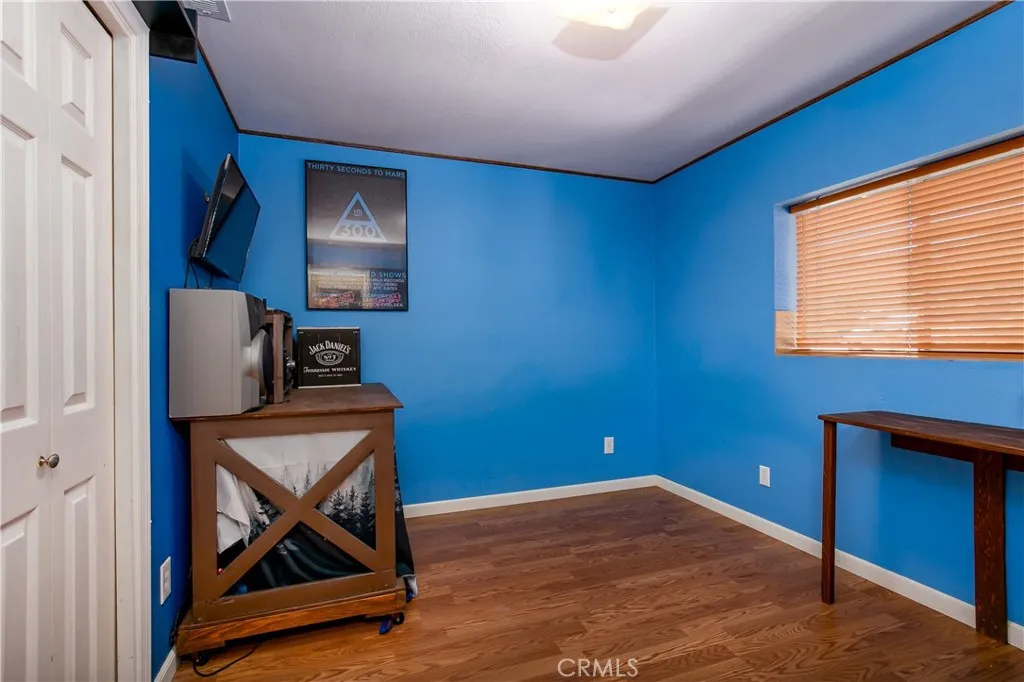 A spacious third bedroom located upstairs off the main hallway. This vibrant room showcases striking blue walls, continuous rich wood flooring, a decorative corner media console setup, and a wide window with wooden blinds that provides excellent natural light.