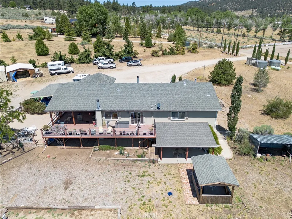 An elevated aerial perspective capturing the rear profile of the home, showcasing the spacious multi-level deck, a detached covered shelter, and the surrounding terrain with mature trees and property parking in the background.