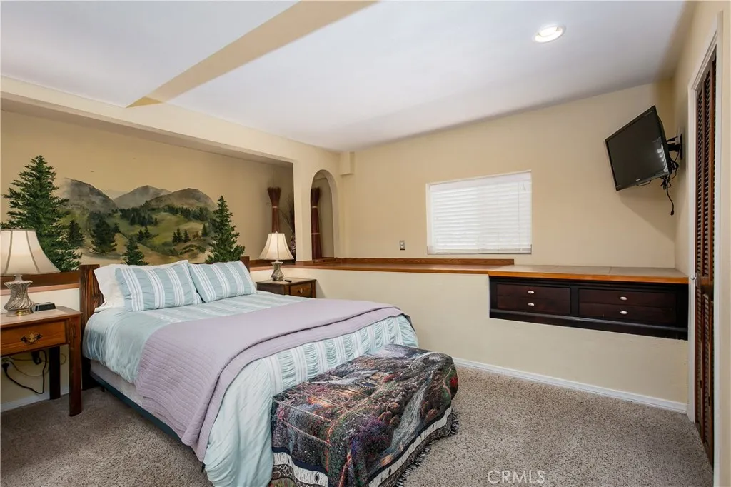 The cozy 5th bedroom, conveniently located on the lower level. This comfortable space features soft carpeting, a unique custom-painted mountain landscape wall mural, a window offering natural light, and a long built-in dark wood dresser with extensive counter space and storage.