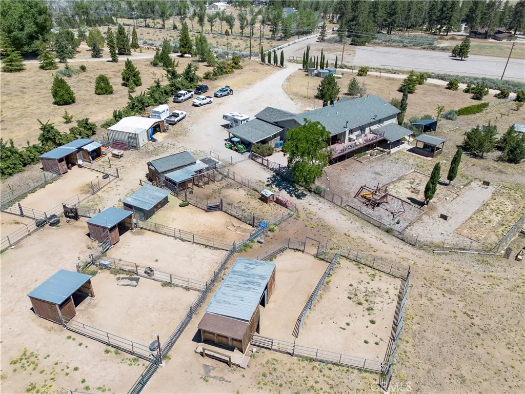 A high-angle aerial view looking from the south, showcasing the rear profile of the home and its expansive deck, alongside the nearby horse shelters, fenced corrals, and a paved driveway leading into the property grounds.