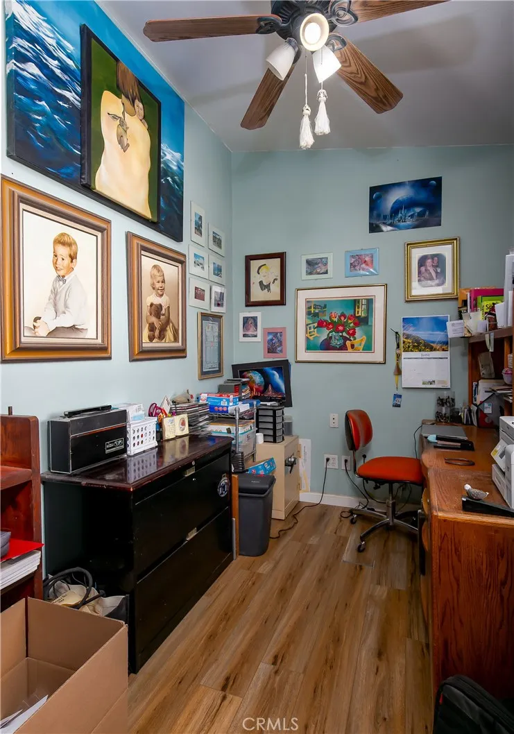 A versatile home office or creative studio space. This practical room features a large wooden desk with ample storage cabinetry, light blue walls, a classic wood-paddle ceiling fan, and durable flooring, offering a perfect private work-from-home setup.
