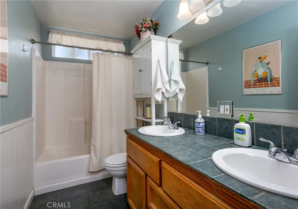 Conveniently located upstairs near the secondary bedrooms, this full hallway bathroom offers a long oak vanity with dual sinks, classic fixtures, and custom teal tile countertops. The space includes a clean shower/tub combination with a white curtain, a standard commode, and a soft blue-grey accent wall.