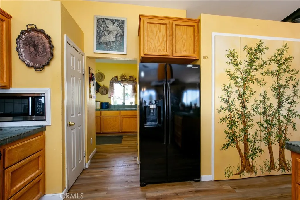 A functional interior perspective showcasing the transition from the kitchen to the utility spaces. This shot highlights a large black refrigerator nestled against warm yellow cabinetry, leading down the hallway toward the laundry room entry, complemented by a unique painted tree mural on the walk-in pantry door.