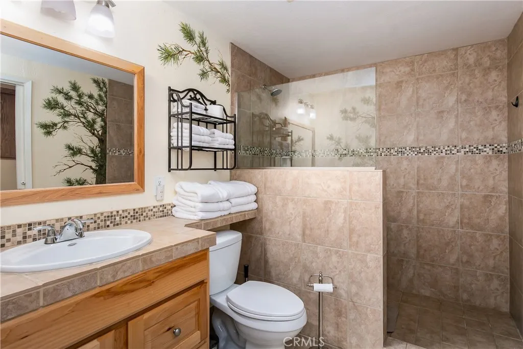 The private en-suite bathroom located inside the downstairs 5th bedroom. This functional space features a warm oak vanity with a matching neutral countertop, a large framed wall mirror, and a spacious walk-in shower lined with large-format floor-to-ceiling tile and a decorative mosaic border.