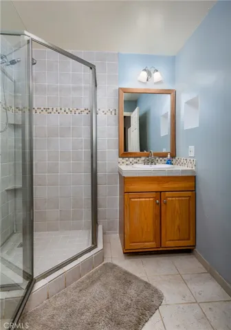 A full 4th bathroom located in the private downstairs suite. This space offers an oak vanity with storage, a large mirror with classic lighting, light blue walls, and a convenient glass-enclosed walk-in shower accented with a mosaic tile border.