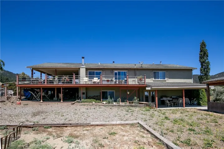 A clear ground-level perspective of the home's rear exterior. This shot highlights the expansive upper-level wooden deck perfect for entertaining, a shaded lower patio area, and the open natural yard space set against a bright, clear blue sky.