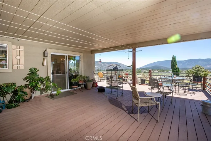 An impressive outdoor living space perfect for entertaining. This large, covered wooden deck features a spacious layout with room for multiple seating arrangements, a dining area, and potted plants, all framed by a sturdy railing overlooking stunning, panoramic mountain and valley views.