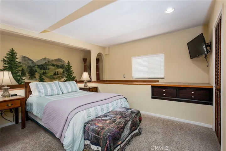 The cozy 5th bedroom, conveniently located on the lower level. This comfortable space features soft carpeting, a unique custom-painted mountain landscape wall mural, a window offering natural light, and a long built-in dark wood dresser with extensive counter space and storage.