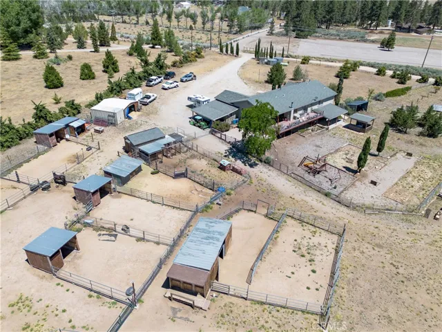 A high-angle aerial view looking from the south, showcasing the rear profile of the home and its expansive deck, alongside the nearby horse shelters, fenced corrals, and a paved driveway leading into the property grounds.