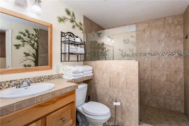 The private en-suite bathroom located inside the downstairs 5th bedroom. This functional space features a warm oak vanity with a matching neutral countertop, a large framed wall mirror, and a spacious walk-in shower lined with large-format floor-to-ceiling tile and a decorative mosaic border.