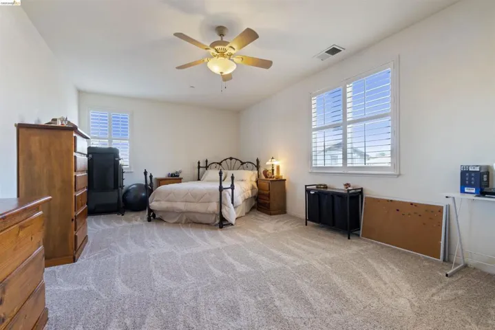 Spacious room featuring carpet flooring, two windows with plantation shutters, a ceiling fan with integrated lighting, and white walls