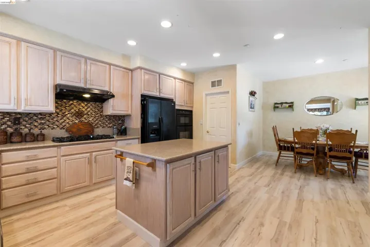 Expansive kitchen featuring light wood-finish cabinetry, a large center island, black appliances, a gas range with an overhead vent hood, and a mosaic tile backsplash
