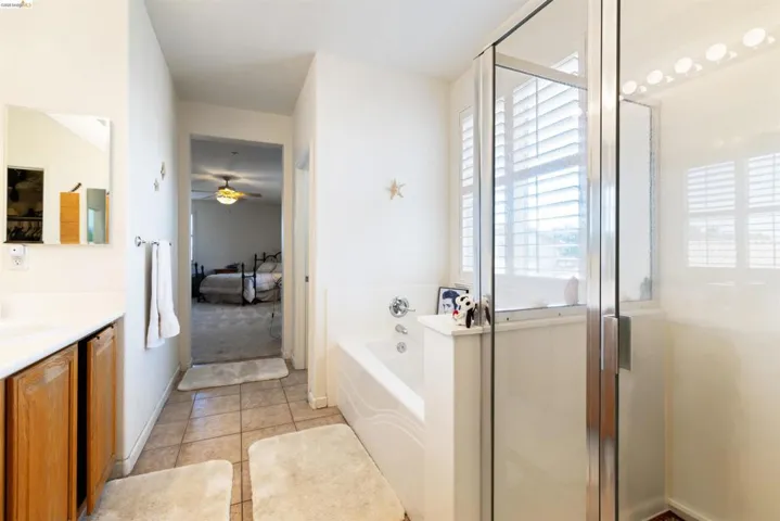Bathroom featuring a glass-enclosed shower, a separate soaking tub with window shutters, a single vanity with wood-finish cabinetry, a framed mirror, and tile flooring