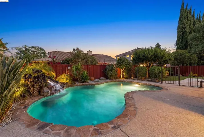Custom-shaped swimming pool featuring a rock waterfall, flagstone coping, integrated lighting, and surrounding concrete decking
