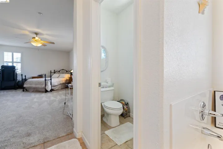 Carpeted room with a ceiling fan, shuttered window, and a white interior door opening to a tiled bathroom with a toilet and oval mirror