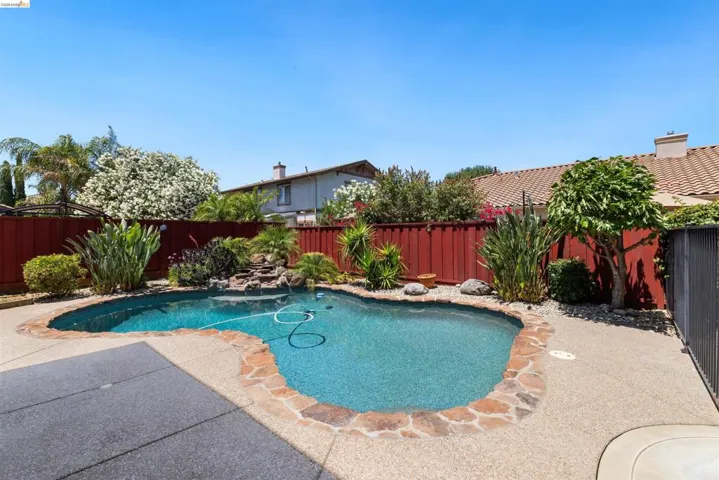 Kidney-shaped swimming pool featuring a stone waterfall, surrounded by lush landscaping, an exposed aggregate deck, and a vibrant red fence