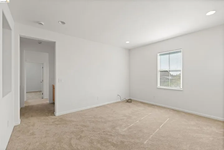 Carpeted interior room featuring a single window, white walls, recessed ceiling lighting, and a doorway leading to a hallway with additional rooms