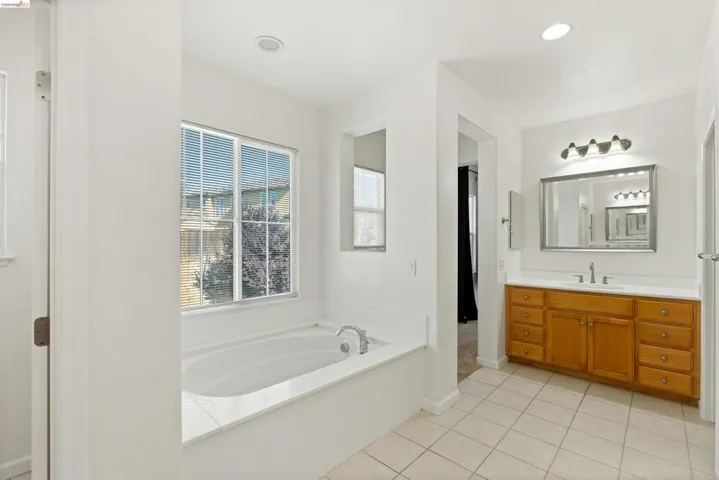 Recessed soaking tub with window, tile flooring, and wood-finish vanity with countertop and integrated sink