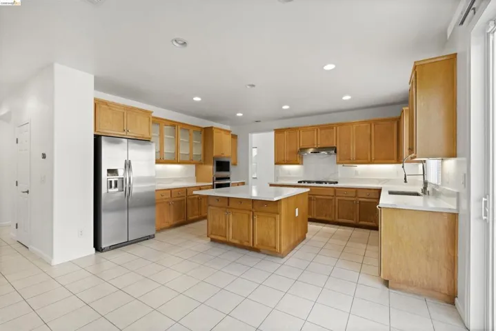 Open-concept kitchen featuring extensive light wood cabinetry, a central island with a white countertop, stainless steel appliances, recessed ceiling lighting, and light-toned tile flooring