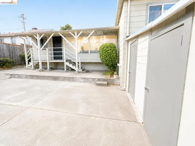 Concrete patio area featuring a covered porch with white railing, exterior staircase, and multiple exterior storage units