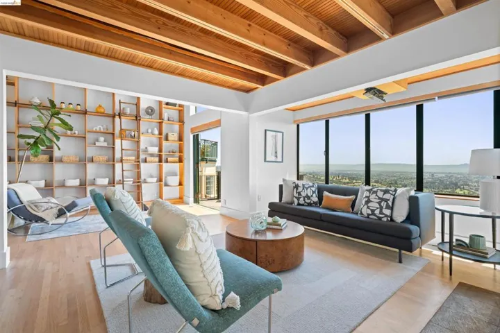 Open-concept living area featuring an exposed beam ceiling, a wall of built-in shelving with a rolling ladder, expansive floor-to-ceiling windows, and light wood-finish flooring