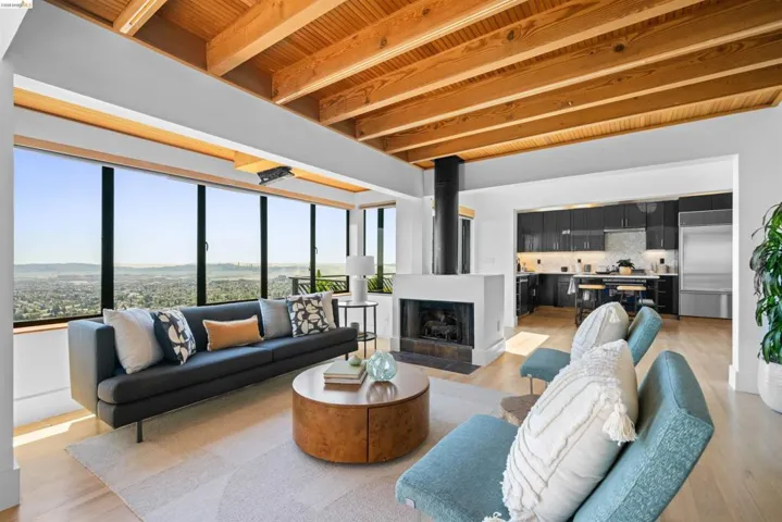 Open-concept living area featuring a wood-beam ceiling, full-height windows, a central fireplace, light wood-finish flooring, and a contemporary kitchen with dark cabinetry and stainless steel appliances