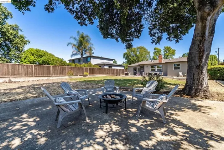 Spacious backyard featuring a concrete patio with a built-in fire pit, mature shade trees, a manicured lawn, and a wooden privacy fence