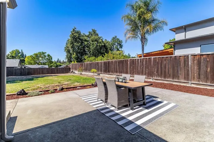 Spacious concrete patio featuring a wood privacy fence, expansive lawn area, mature trees, and a modern exterior building with multiple windows