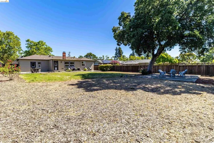 Expansive backyard featuring a large mature tree, wood chip landscaping, a private wooden fence, a concrete patio, and a stucco exterior with a brick chimney