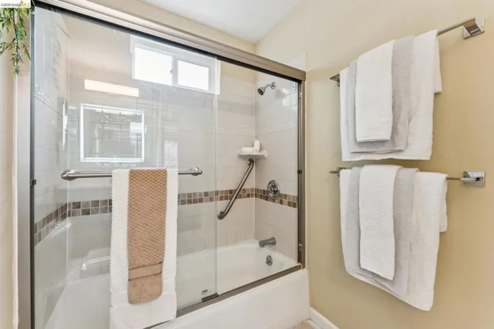 Bathtub with a glass sliding door enclosure, tiled surround featuring a decorative mosaic border, and a grab bar