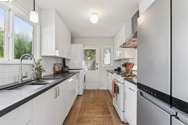 Galley kitchen featuring white cabinetry, dark countertops, a stainless steel refrigerator, a gas range with a stainless steel hood, and a dual basin sink with a gooseneck faucet