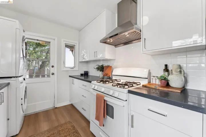 Modern kitchen featuring white cabinetry, dark countertops, a gas range with stainless steel hood, and a stacked washer and dryer unit