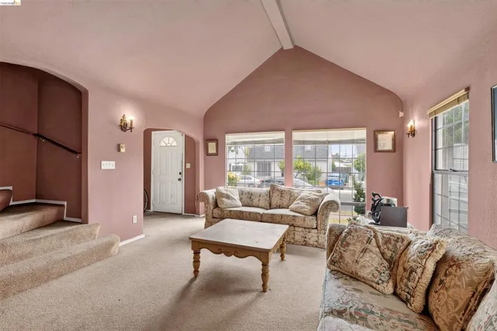 Living area featuring a vaulted ceiling with exposed beam, multiple large windows, wall sconces, and a white paneled front door