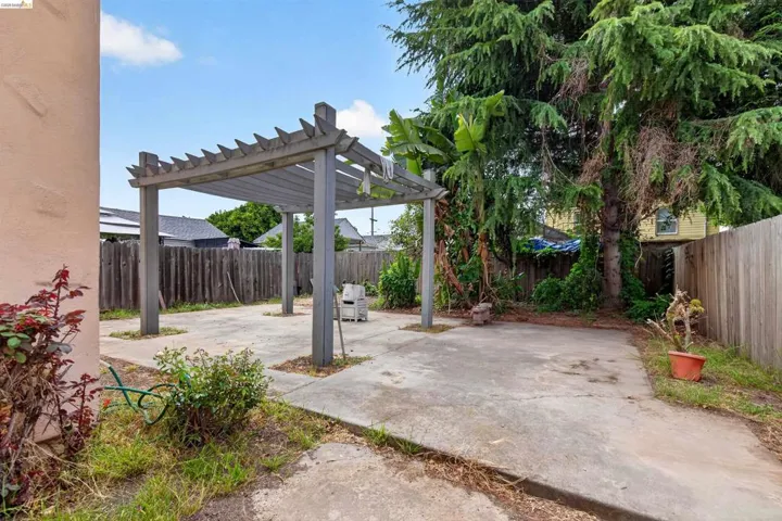 Outdoor patio area featuring a wooden pergola over concrete paving, surrounded by a wood plank fence and mature trees
