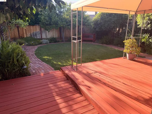 Expansive backyard featuring a red-toned wood deck, a circular grass lawn, a brick-laid pathway, a privacy fence, and mature trees