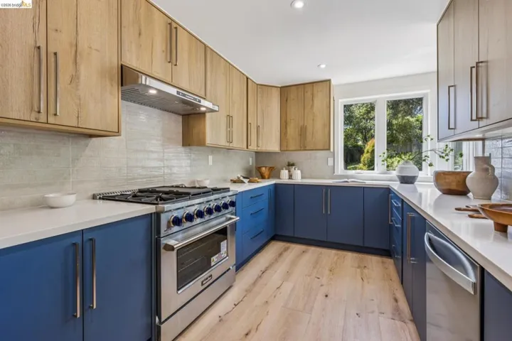 Kitchen featuring two-tone cabinetry with wood-finish upper cabinets and blue lower cabinets, stainless steel range with matching hood, integrated dishwasher, light-toned backsplash, and white countertops