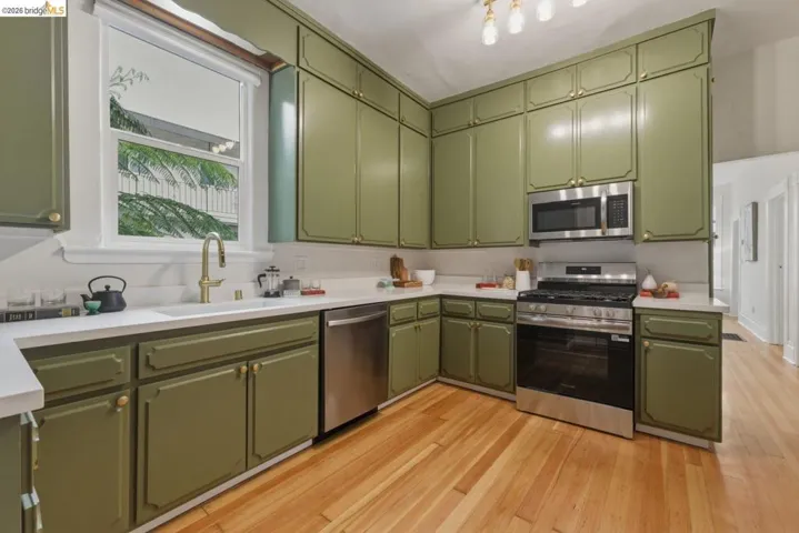 Kitchen featuring abundant olive-toned cabinetry, stainless steel appliances, a white single-basin sink with a brass-finish faucet, light-toned countertops, and wood-finish flooring