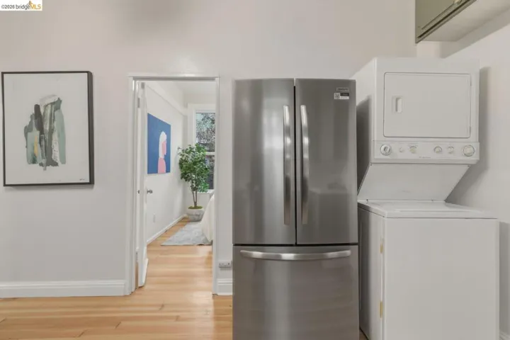 Stainless steel French door refrigerator, stacked washer and dryer, wood-finish flooring, white walls, and a doorway leading to a separate area