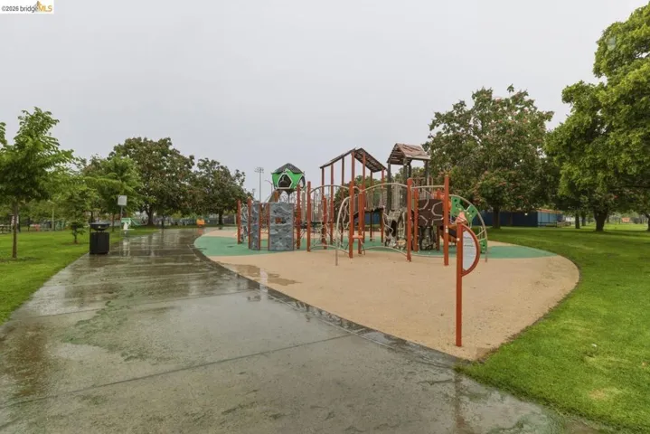 Outdoor playground area featuring multiple structures, sand-colored ground covering, and surrounding green spaces