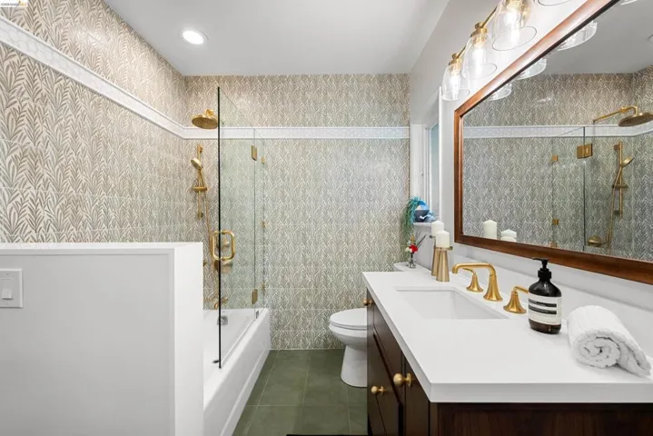 Bathroom featuring a built-in tub with a glass shower enclosure, brass fixtures, a vanity with a white countertop and integrated sink, and patterned wall tile with a decorative border