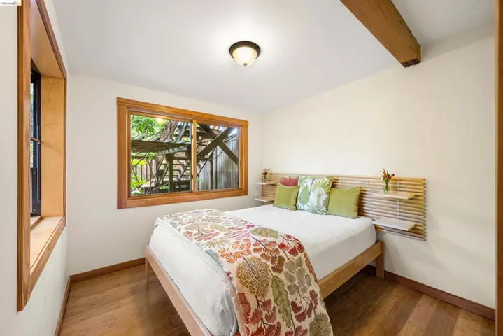 Bedroom featuring wood-finish flooring, two windows with wood trim, a built-in slatted wood headboard with integrated shelving, a ceiling-mounted light fixture, and an exposed wood beam
